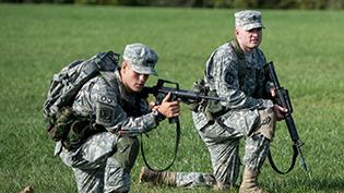 Two men in army uniforms crouch and practice firing their weapons.